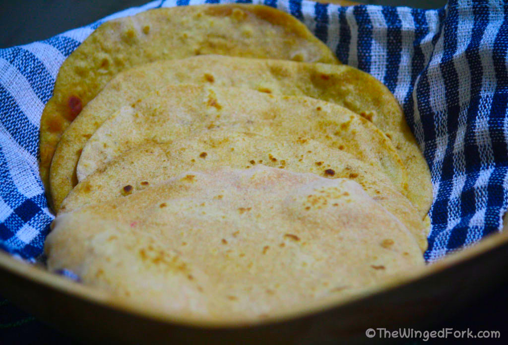 Multi-grain chappatis ready to be served in a wicker basket on top of a blue and white checquered towel.
