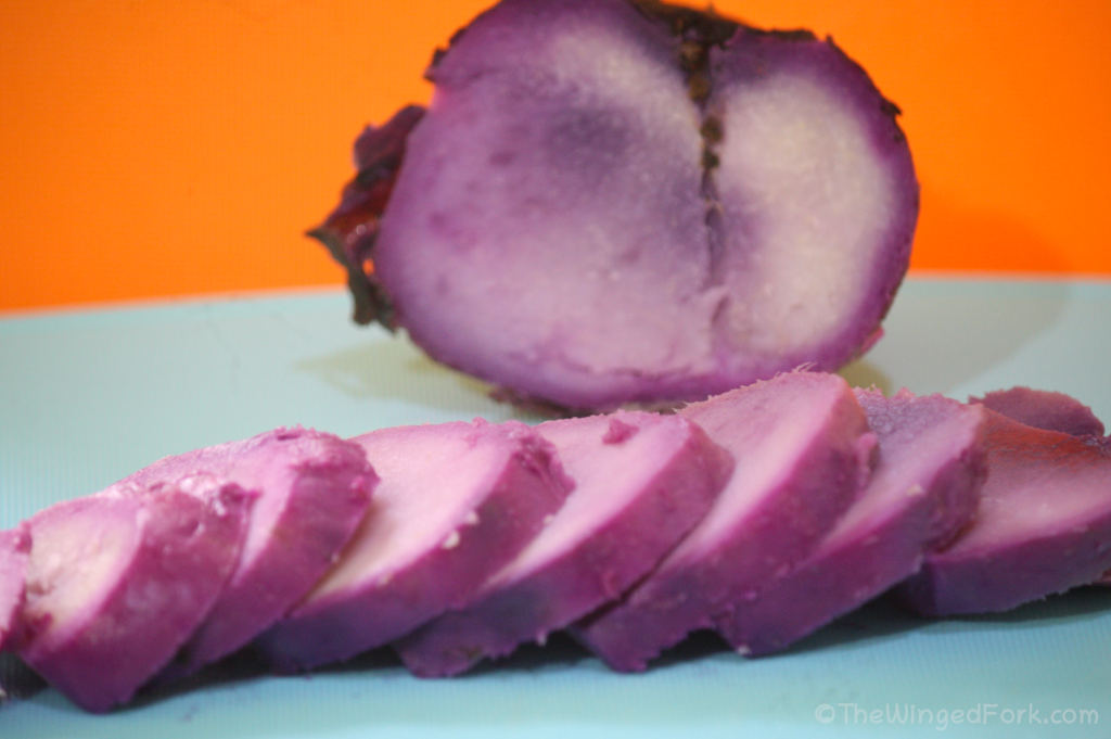 Slices of purple yam in front of a half piece on a blue cutting board and orange background.