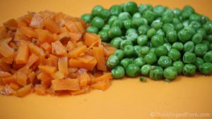 Bolied carrot pieces and green peas on a orange cutting board.