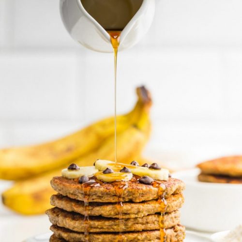 Banana pancakes topped with banana slices and chocolated chips and a jar of syrup with background bananas.