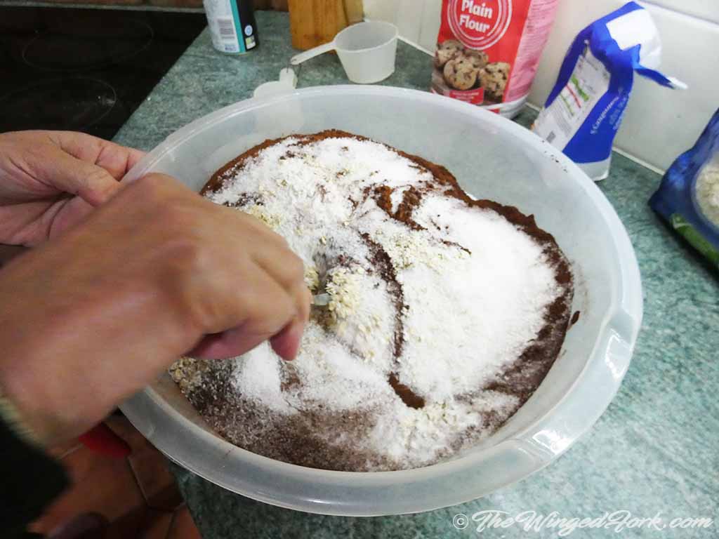 Desiccated coconut, cocoa and oats being mixed with a spoon in a bowl.