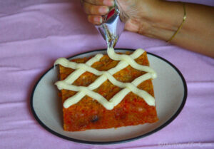 A hand decorating a carrot cake with cream cheese frosting.