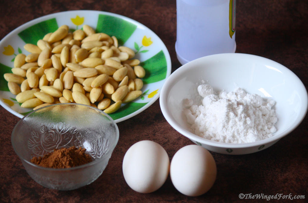 Almonds, eggs, powdered sugar, cocoa powder in bowls and a bottle on a brown platform.