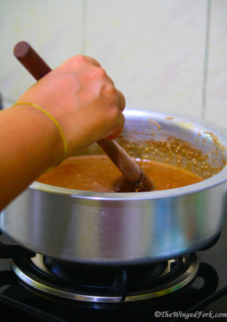 A hand holding a wooden spoon stirring a mixture in a vessel on the stove.